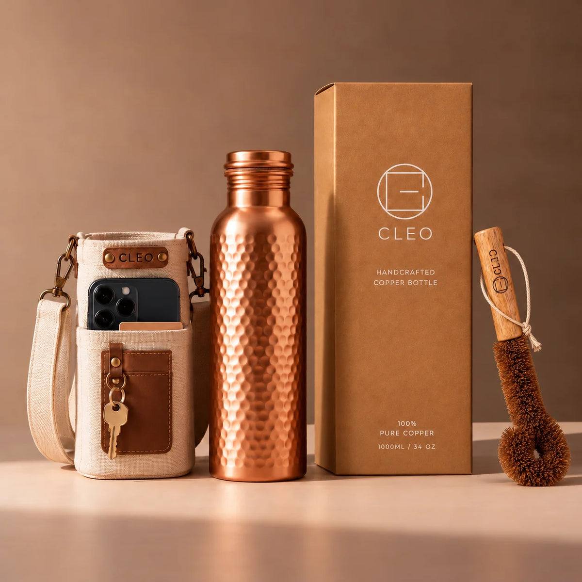 CLEO brand set featuring a hammered copper water bottle, a cream and brown phone pouch with a phone inside, and a brown cleaning brush, arranged next to their kraft paper packaging boxes on a warm beige background.